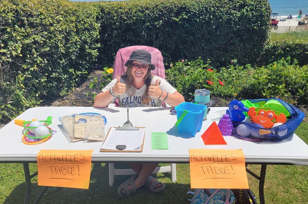 Woman sitting at a table outdoors with childrens toys in baskets on the top of the table and an orange sign that says Activities Table