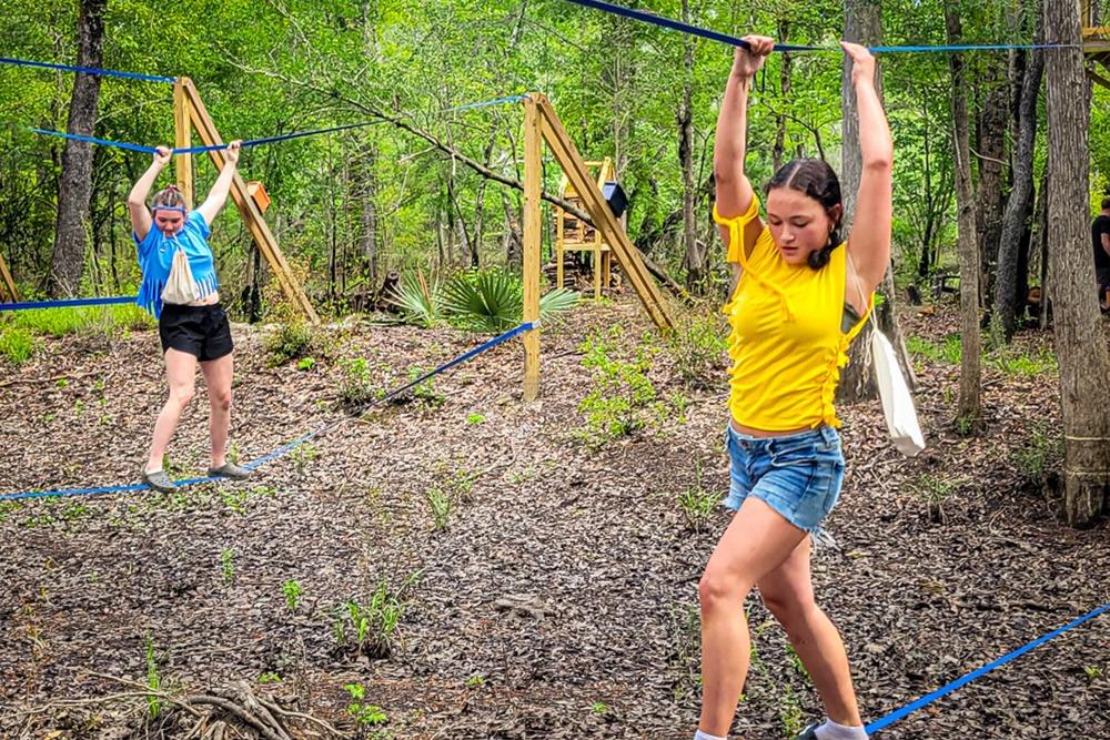Two girls are engaging in an outdoor rope course challenge in a wooded area. The girl in the foreground, wearing a yellow t-shirt and denim shorts, is carefully navigating a balance element, holding onto blue ropes above her head for support.