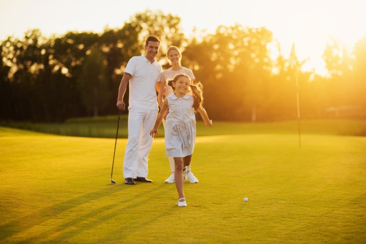 A man with a woman stands next to each other with golf clubs in their hands, the girl in front of them runs toward the camera and smiles