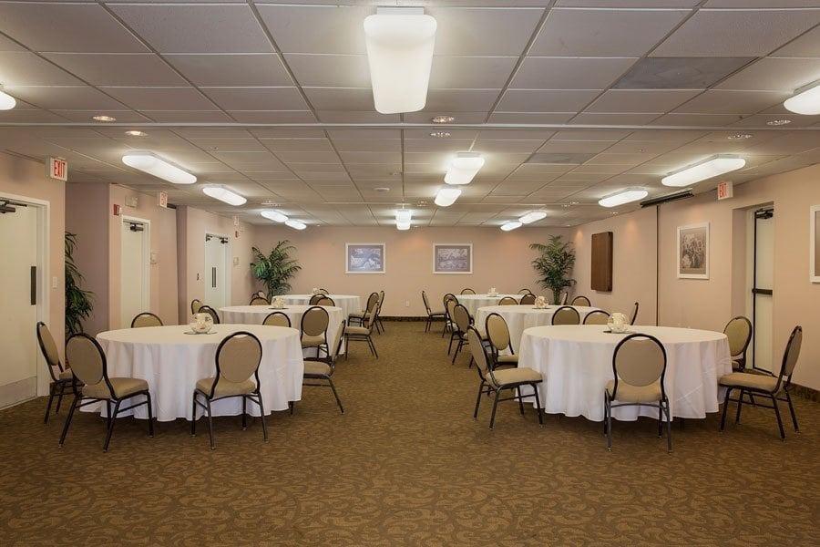 An indoor banquet hall with multiple round tables covered with white tablecloths, surrounded by padded chairs with metal frames.