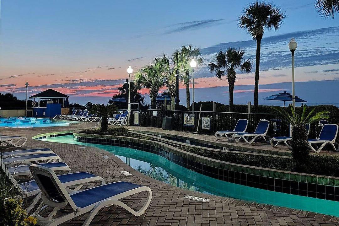 Twilight at Grande Shores with poolside loungers, palms, and a colorful sky.