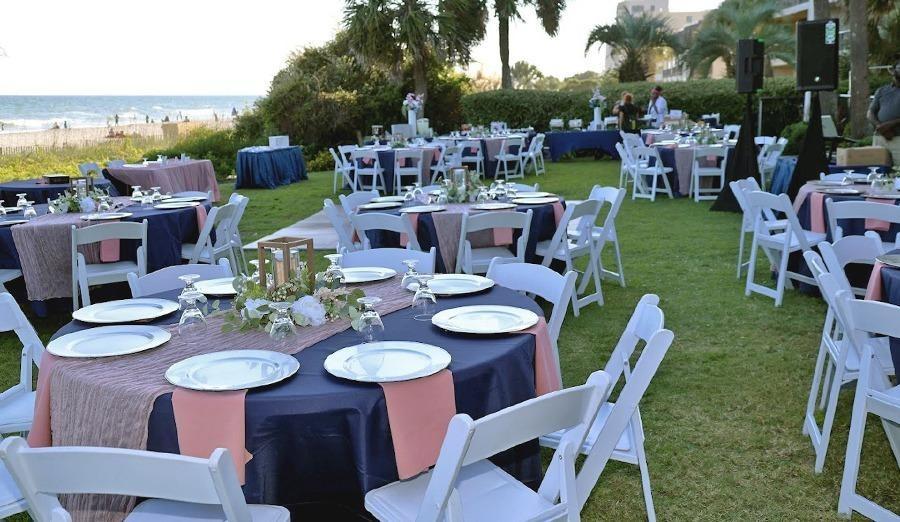 An outdoor wedding setup with rows of empty white chairs facing a wooden arch decorated with purple ribbons and white and purple flowers.