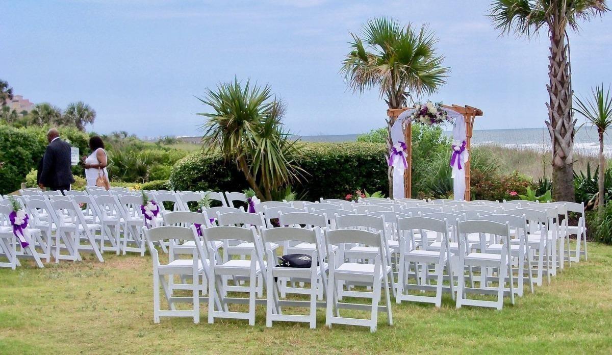 An outdoor wedding setup with rows of empty white chairs facing a wooden arch decorated with purple ribbons and white and purple flowers.