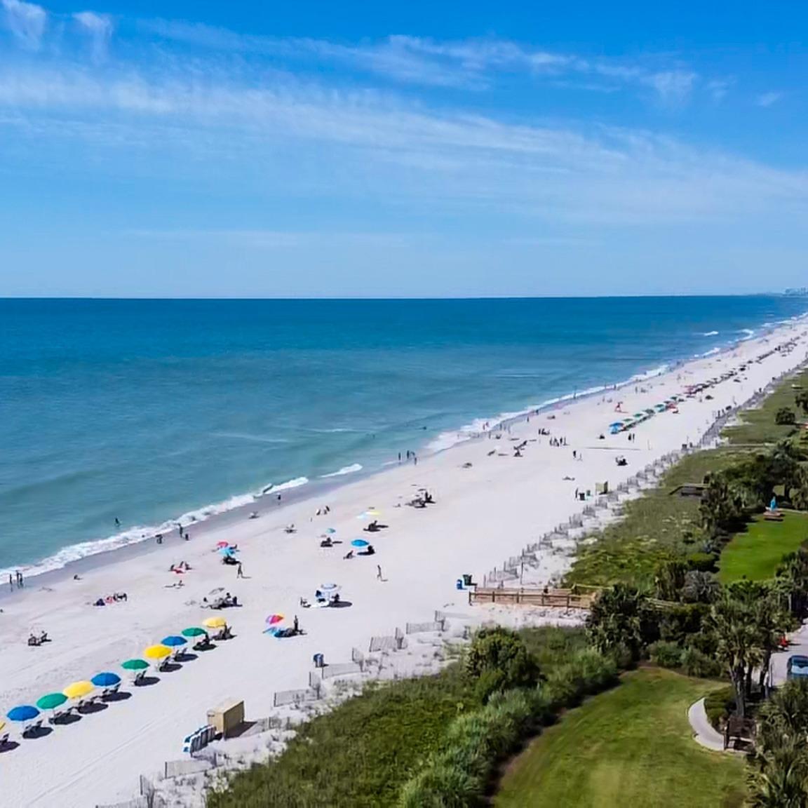 photo of the beach in front of Grande Shores Resort showing the sand, beachgoers, and the ocean with a blue sky with clouds