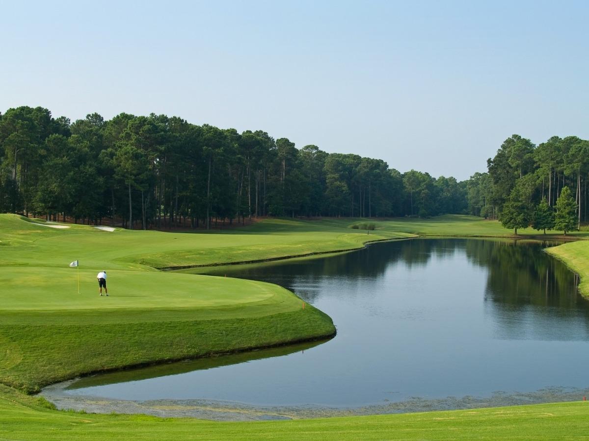 Scenic view of the Myrtle Beach Classic PGA Tour golf course, featuring a wide pond curving alongside the green and a golfer in a white shirt preparing to putt near the flag, with tall pine trees lining the background.