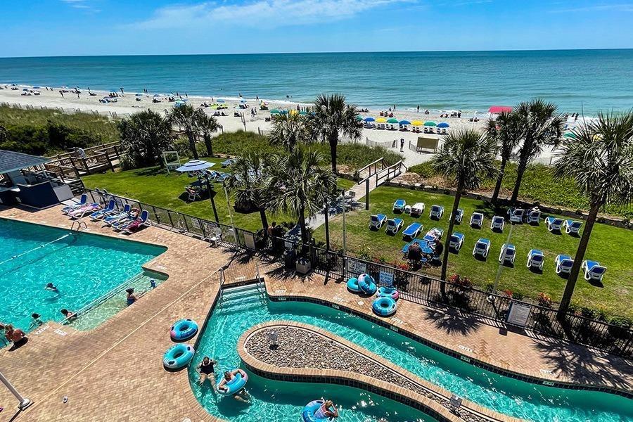 Aerial view of Grande Shores Ocean Resort's oceanfront pool area, featuring a lazy river, swimming pool, lounge chairs, direct access to the beach, and stunning ocean views.