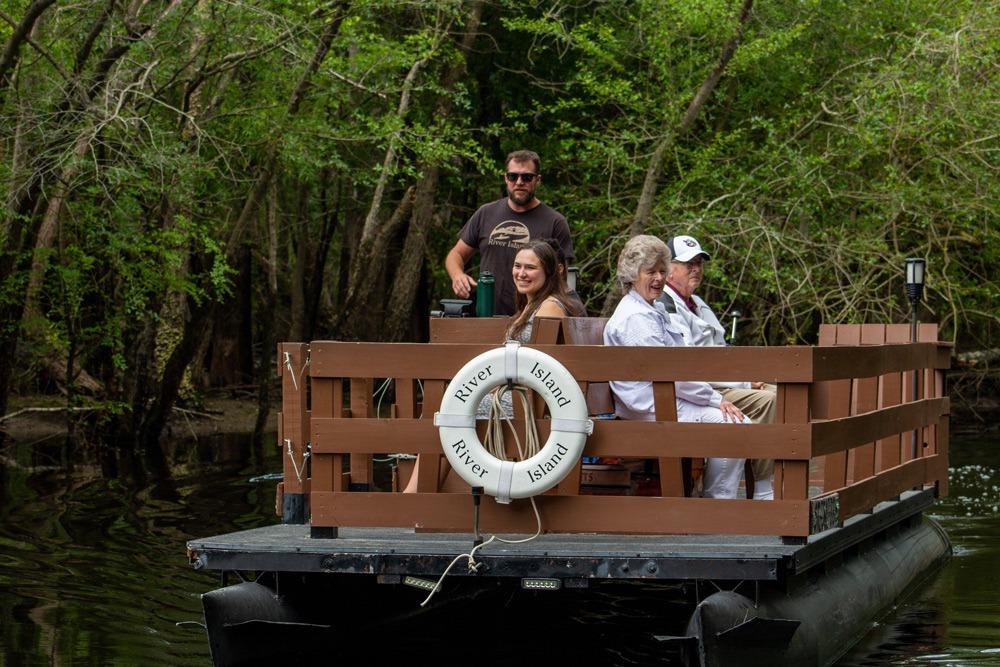 Three passengers and a boat operator are aboard a small, flat-bottomed riverboat adorned with life rings labeled 'River Island