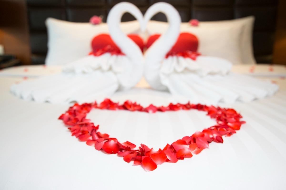 Two swans (made from towels) form a heart on a hotel suite bed, with flowers surrounding them.